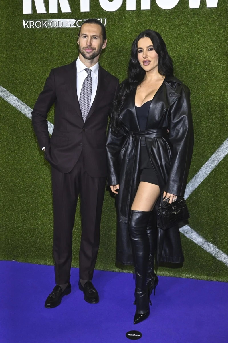 Elegancka para pozuje na niebieskim dywanie przed zieloną ścianą z napisem, mężczyzna ubrany w garnitur, kobieta w czarnych kozakach, krótkiej sukience oraz długim płaszczu.