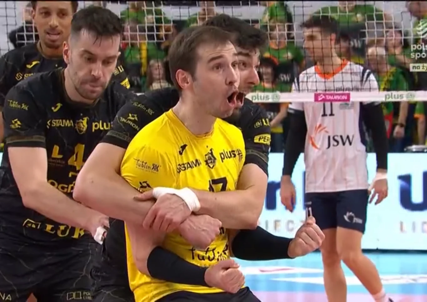 Grupa siatkarzy w żółto-czarnych strojach świętujących punkt na boisku, jeden zawodnik w centrum wyraża silne emocje z szeroko otwartymi ustami, w tle widoczni inni sportowcy oraz siatka.