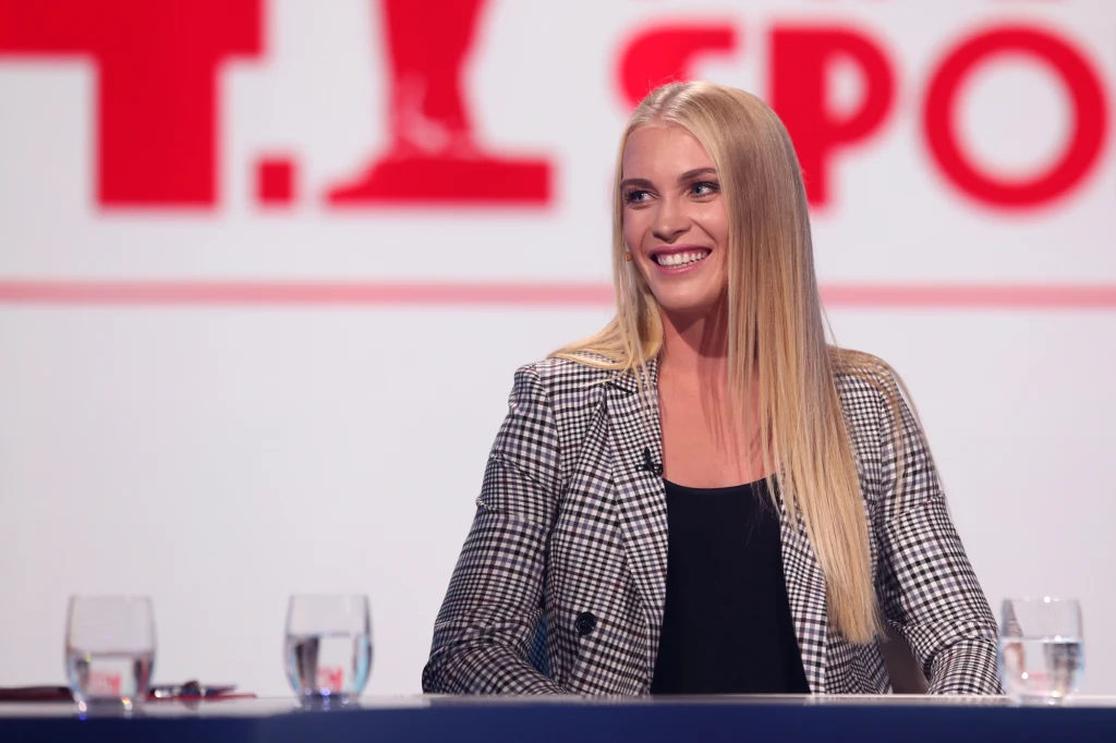 Kobieta o blond włosach w marynarce w kratę siedzi przy stole w studiu telewizyjnym, przed nią stoją trzy szklanki z wodą, w tle rozmazany napis sugerujący kanał sportowy.