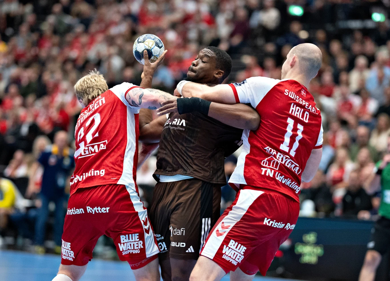 Dylan Nahi w Aalborgu Dynamiczna walka podczas meczu piłki ręcznej, zawodnik w ciemnym stroju próbuje rzucić piłkę, blokowany energicznie przez dwóch przeciwników w czerwono-białych strojach, na tle zapełnionych kibicami trybun.