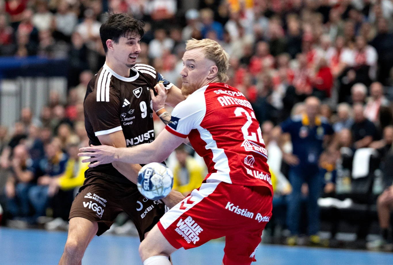 Szymon Sićko Dwóch zawodników piłki ręcznej walczących o piłkę na boisku, otoczonych przez skupioną publiczność na trybunach, jeden z graczy w czerwono-białym stroju, drugi w brązowo-białym.