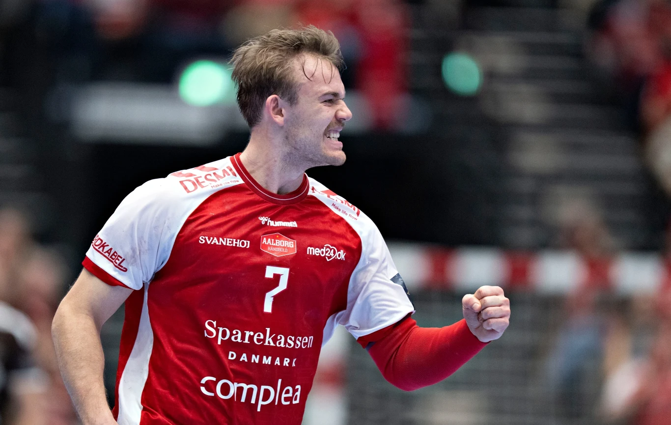 Thomas Arnoldsen Zawodnik drużyny piłki ręcznej ubrany w czerwono-biały strój z numerem 7 na piersi, okazujący radość i triumf, zaciśnięta pięść, tło rozmyte z widocznymi kibicami na trybunach.