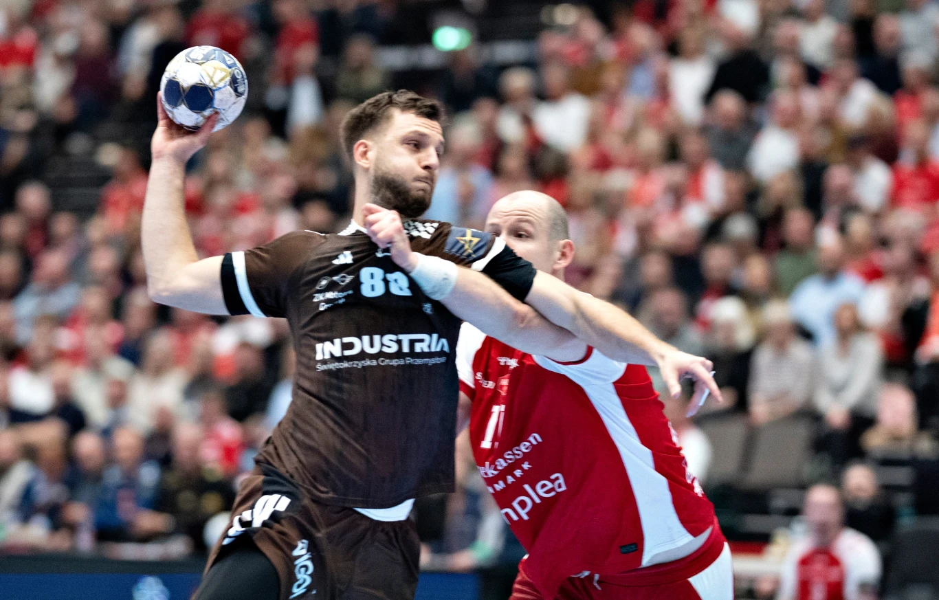 Aleks Vlah wrócił do Aalborga - tym razem jako zawodnik Industrii Dwóch zawodników w dynamicznej walce o piłkę ręczną podczas meczu na hali sportowej, trybuny wypełnione kibicami, zawodnik w brązowej koszulce przygotowuje się do oddania rzutu, nad nim unosi się piłka.