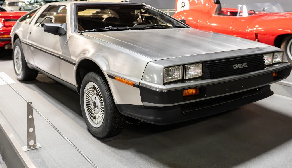 Srebrny samochód DeLorean DMC-12 z charakterystyczną, futurystyczną sylwetką oraz drzwiami unoszonymi do góry stoi na ekspozycji w muzeum motoryzacji, w otoczeniu innych klasycznych pojazdów.