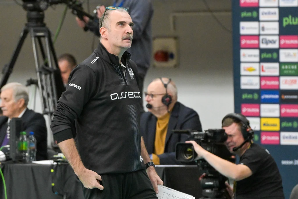 Massimo Botti, trener Asseco Resovii Trener drużyny sportowej ubrany w czarną bluzę z logiem sponsora stoi przy linii bocznej hali sportowej, trzymając kartkę i spoglądając na boisko; w tle operatorzy kamer oraz osoby komentujące wydarzenie.