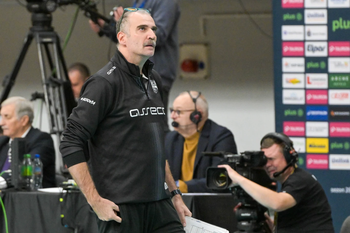 Trener drużyny sportowej ubrany w czarną bluzę z logiem sponsora stoi przy linii bocznej hali sportowej, trzymając kartkę i spoglądając na boisko; w tle operatorzy kamer oraz osoby komentujące wydarzenie.