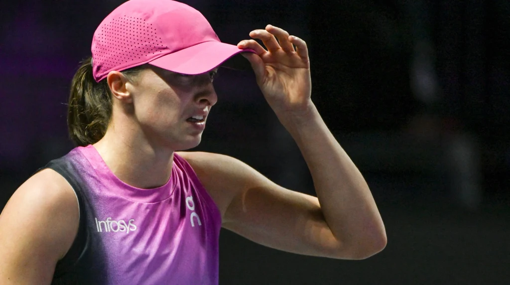 Iga Świątek Kobieta w stroju sportowym oraz różowej czapce z daszkiem, podczas intensywnego wysiłku fizycznego, w jasno oświetlonym miejscu, zyskująca skupiony wyraz twarzy.