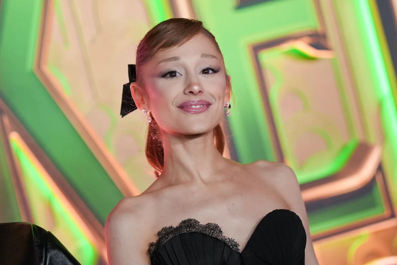 Ariana Grande porzuca muzykę na rzecz aktorstwa? Młoda kobieta w eleganckiej czarnej sukni z koronkowym wykończeniem, uczesane włosy spięte z tyłu, subtelny makijaż. W tle widoczne kolorowe dekoracje w zielono-złotych barwach.