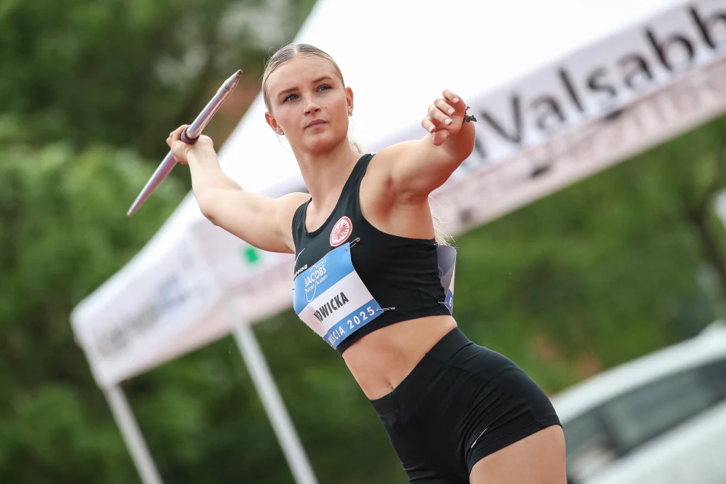 Zawodniczka przygotowująca się do rzutu oszczepem, ubrana w strój sportowy z numerem startowym, skupiona, w tle rozmyte elementy stadionu i baner reklamowy.