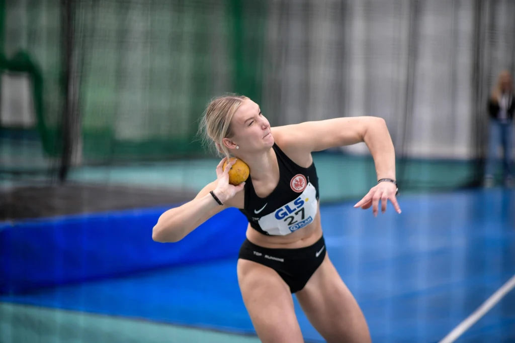Zawodniczka w sportowym stroju przygotowuje się do pchnięcia kulą na hali lekkoatletycznej, skupiona na technice i ruchu. W tle niebieska mata oraz rozmazana siatka zabezpieczająca.