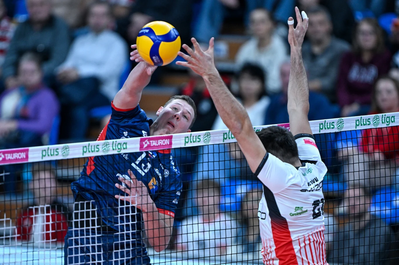 Dwóch zawodników rywalizuje podczas meczu siatkówki, jeden z nich odbija piłkę ponad siatką, próbując zdobyć punkt, a drugi gracz usiłuje zablokować atak. W tle widoczna publiczność oraz trybuny.