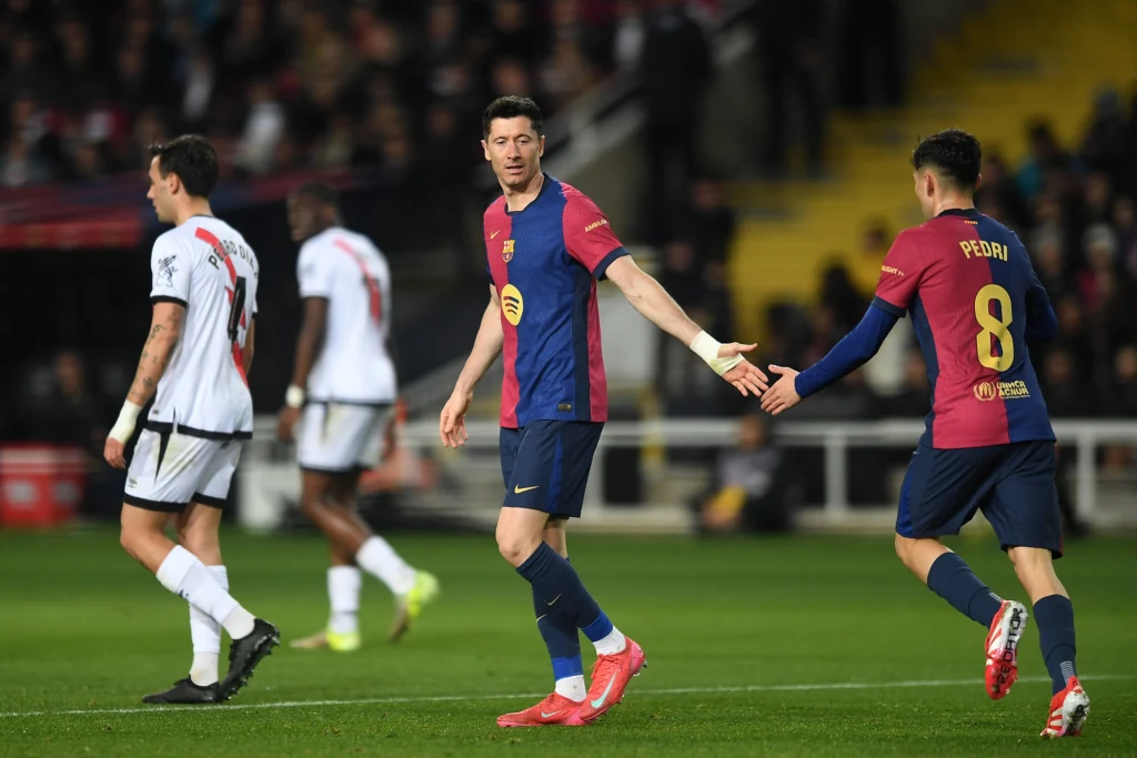 Zawodnik FC Barcelony wyciąga rękę do kolegi z drużyny podczas meczu piłkarskiego, w tle piłkarze drużyny przeciwnej i trybuny z kibicami.