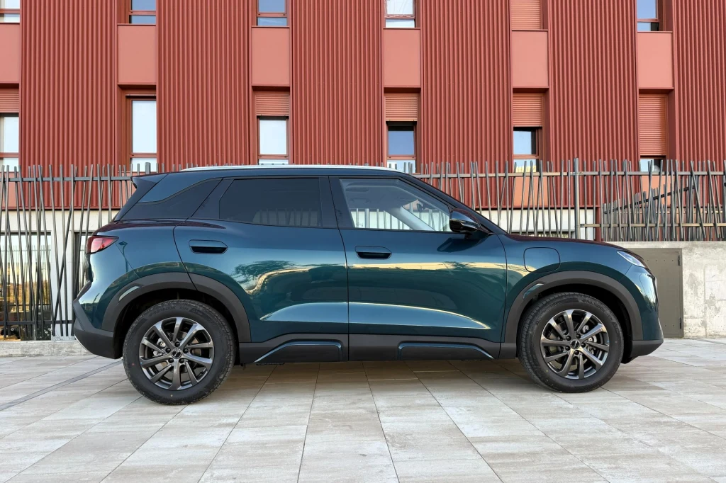 Nowoczesny zielony SUV na tle czerwonego budynku z wertykalnymi oknami oraz metalowym ogrodzeniem, zaparkowany na jasnej, kamiennej nawierzchni.