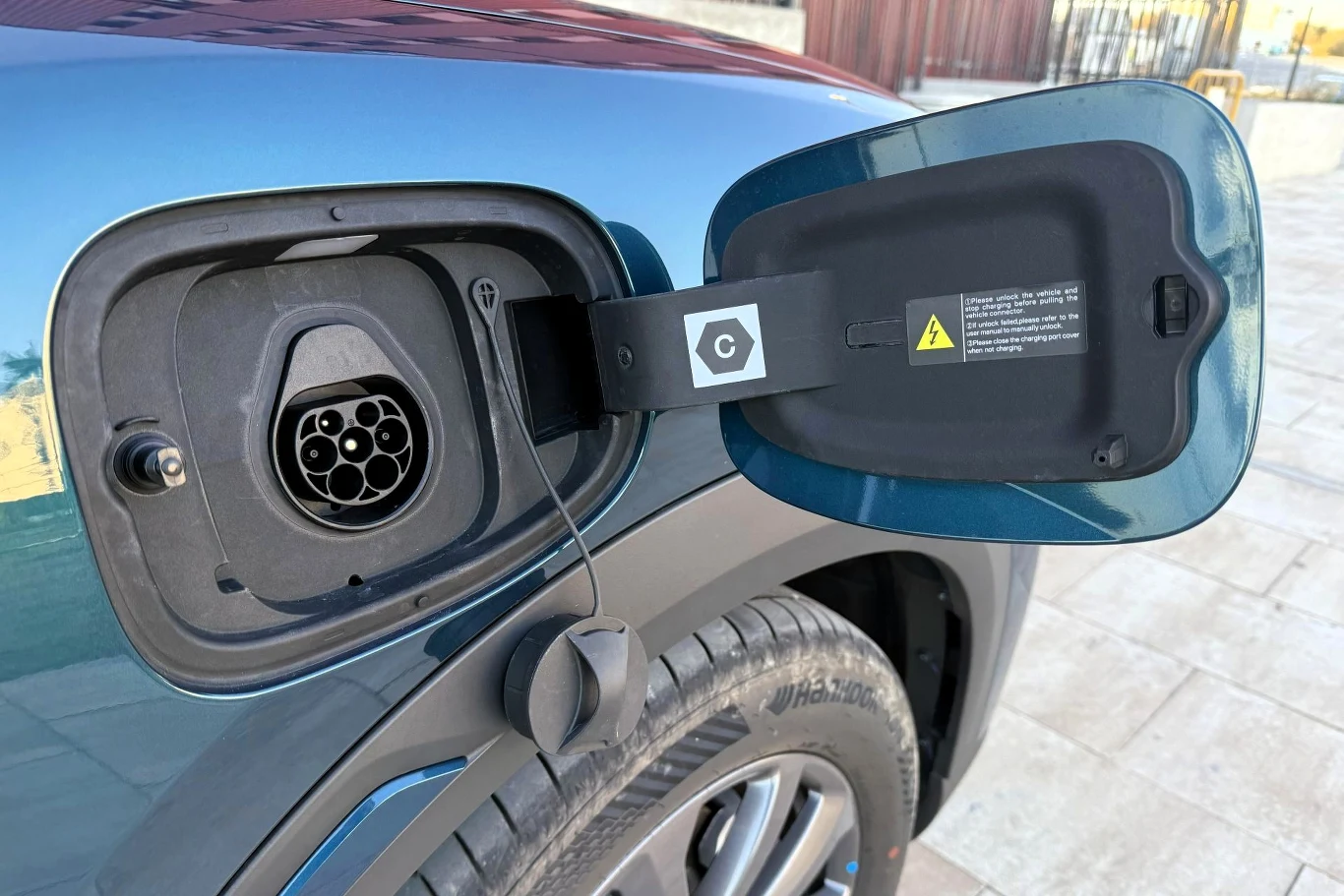 W obu wariantach mocy BYD Atto 2 DM-i obsługuje tylko ładowanie prądem przemiennym. Otwarty wlew do ładowania samochodu elektrycznego z widocznym gniazdem ładowania oraz fragmentem tylnego koła pojazdu, pokryty metalicznym lakierem. Pokrywa wlewu otwarta, a na niej widoczna jest etykieta z ostrzeżeniem i symbolem ładowania.