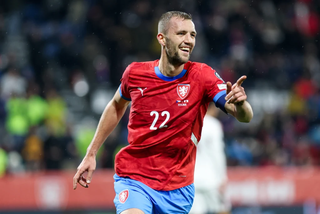 Piłkarz w czerwonej koszulce z numerem 22 świętuje zdobycie bramki podczas meczu piłkarskiego, wskazując palcem w kierunku trybun, na tle rozmazanej publiczności.