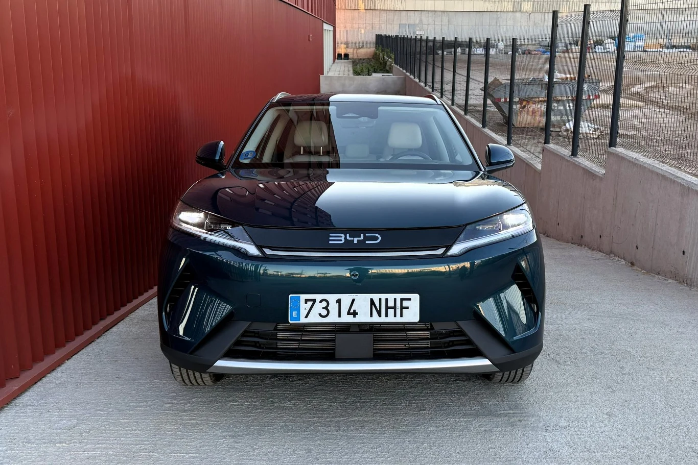 Na pochwałę zasługuje zaskakująco dobre, jak na auto tego segmentu, wyciszenie. Nowoczesny samochód osobowy marki BYD ustawiony frontem na tle czerwonego budynku i ogrodzenia z siatki, z wyraźnie widoczną tablicą rejestracyjną.