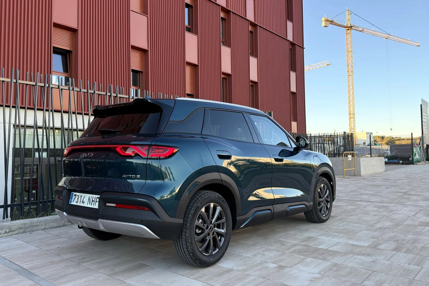 BYD Atto 2 DM-i dostępne jest w dwóch wersjach wyposażonych połączonych z wariantami napędu. SUV w kolorze ciemnozielonym zaparkowany na tle nowoczesnego budynku z czerwonymi panelami, w oddali widoczny żółty żuraw budowlany.