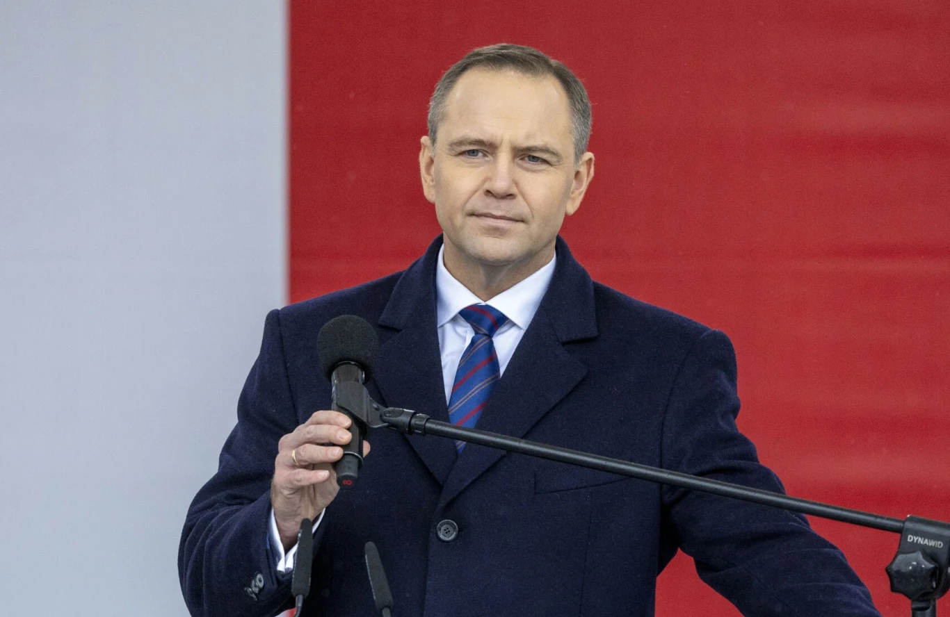 Mężczyzna w granatowym płaszczu i koszuli z krawatem stoi przed mikrofonem na tle fragmentu dużej flagi w barwach czerwono-białych.