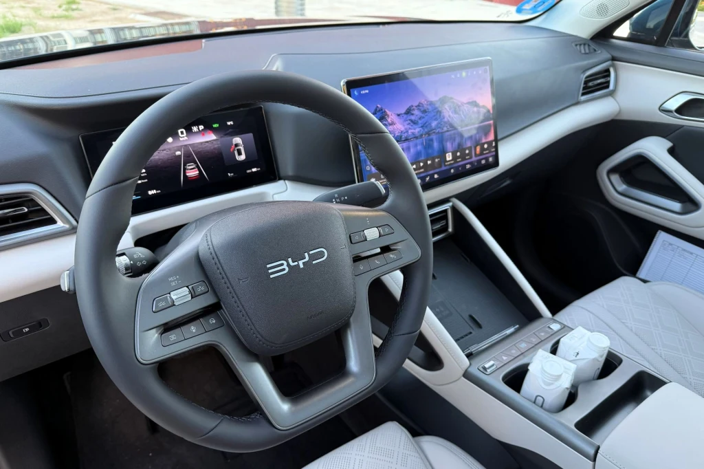 Nowoczesne wnętrze samochodu marki BYD z dużym ekranem dotykowym na konsoli środkowej, cyfrowymi wskaźnikami oraz wielofunkcyjną kierownicą, w jasnej tapicerce.