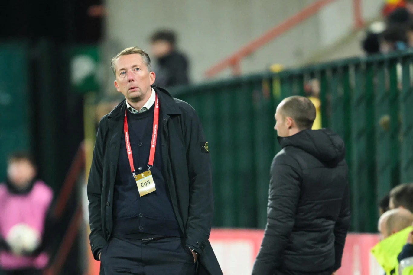 Mężczyzna w ciemnym płaszczu z identyfikatorem na szyi stoi obok innego mężczyzny w kurtce na tle stadionowych barierek, w tle rozmazane postacie związane z wydarzeniem sportowym.