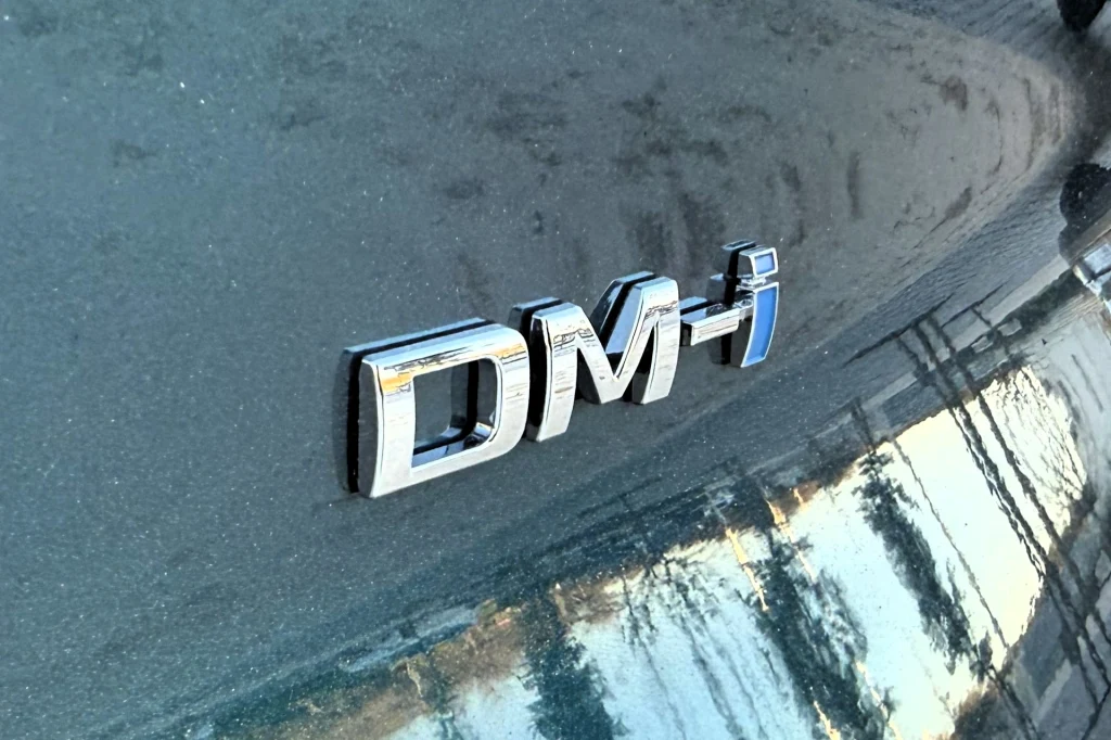 Błyszczący emblemat DM-i umieszczony na metalicznie zielonym nadwoziu samochodu, odbijający światło i pobliskie otoczenie.