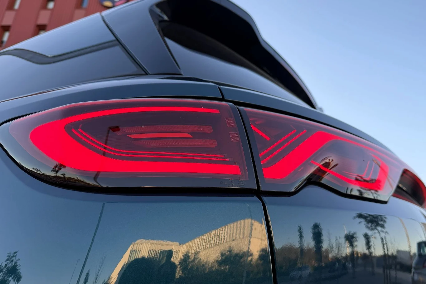 Z zewnątrz stylistyka BYD Atto 2 wciąż nie porywa. Zwracają na siebie uwagę chyba tylko tylne światła przypominające symbol nieskończoności. Zbliżenie na nowoczesną, podświetloną tylną lampę samochodu, na tle budynku i nieba o delikatnym odcieniu błękitu, odbijające się delikatnie na lakierze pojazdu.