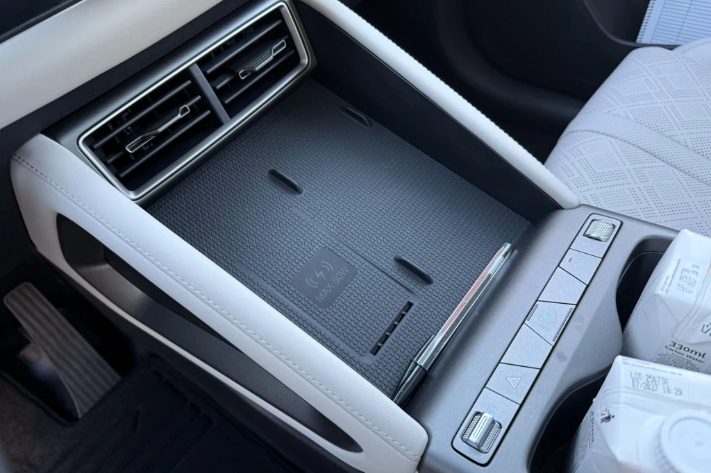 Konsola środkowa samochodu z widocznymi kratkami nawiewu, miejscem na telefon z funkcją ładowania indukcyjnego oraz panelem przycisków sterujących. Na bocznym podłokietniku znajdują się kartoniki z napojami.