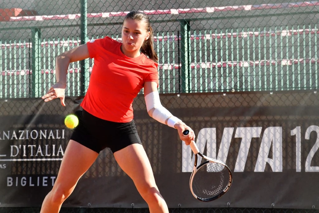 Tenisistka wykonująca uderzenie forhendowe podczas meczu na korcie z czerwoną nawierzchnią. Ubrana w czerwoną koszulkę i czarne szorty, skoncentrowana na piłce zbliżającej się do rakiety. Za zawodniczką widoczne ogrodzenie oraz baner reklamowy.