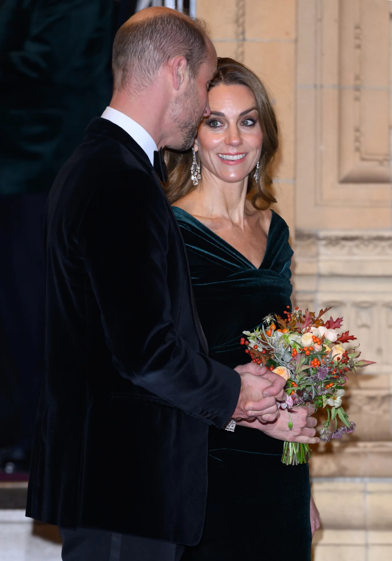 Elegancko ubrana kobieta w zielonej, aksamitnej sukni trzyma kolorowy bukiet kwiatów, stoi obok mężczyzny w czarnym garniturze, spoglądając na niego z ciepłym uśmiechem. Tło stanowi jasna, dekoracyjna ściana.