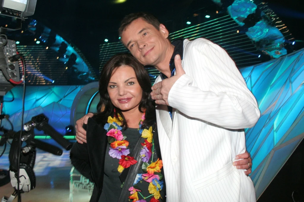 Mężczyzna w białej marynarce obejmuje kobietę z kolorowym naszyjnikiem z kwiatów, oboje uśmiechnięci pozują do zdjęcia na tle jasnych świateł i geometrycznych dekoracji scenicznych.