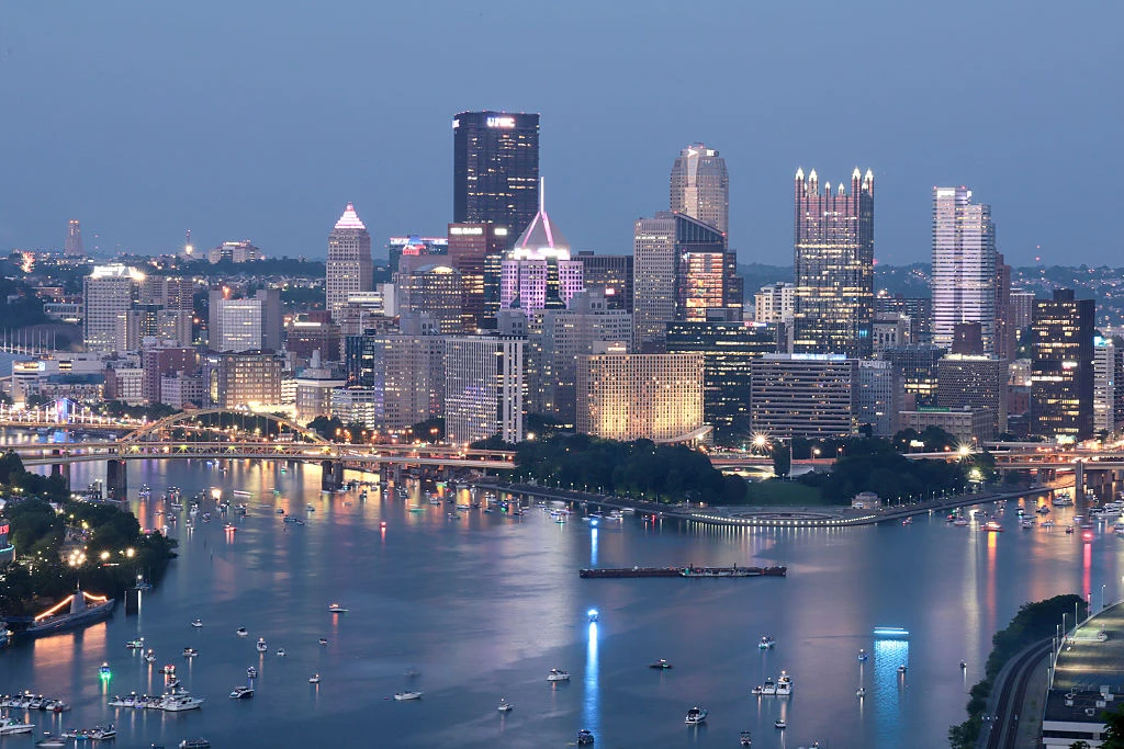 Panorama miasta nocą z wieżowcami oświetlonymi światłami, odbicia w wodzie rzeki, na której unoszą się liczne łodzie i jachty.
