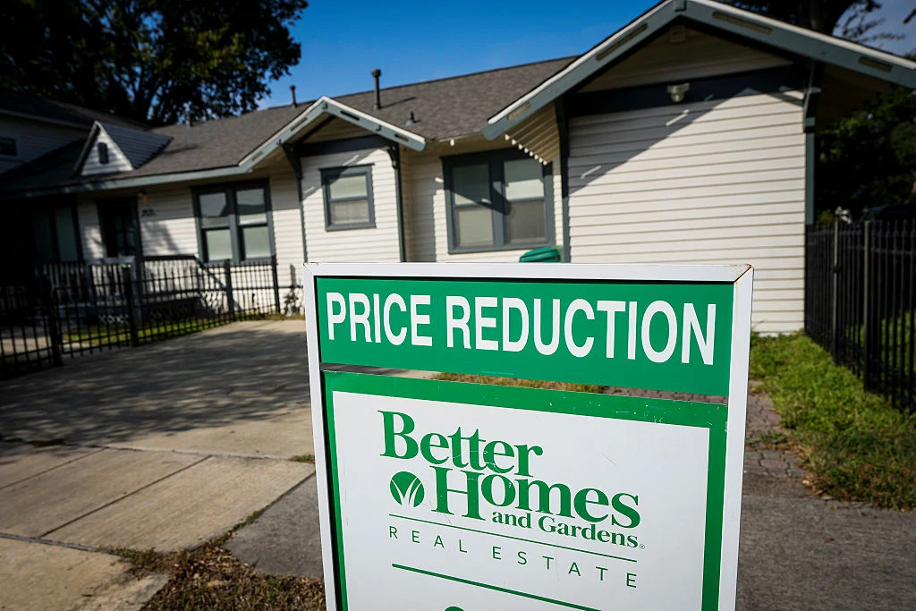 Tablica informująca o obniżce ceny nieruchomości firmy Better Homes and Gardens Real Estate na tle budynku mieszkalnego. Przed domem widoczny jest chodnik oraz ogrodzenie wokół posesji.