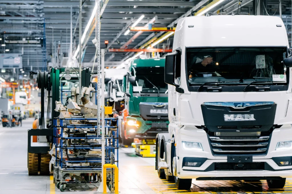 Linia produkcyjna pojazdów ciężarowych w nowoczesnej fabryce z widocznymi ciężarówkami MAN oraz stanowiskami montażowymi i pracującymi maszynami.