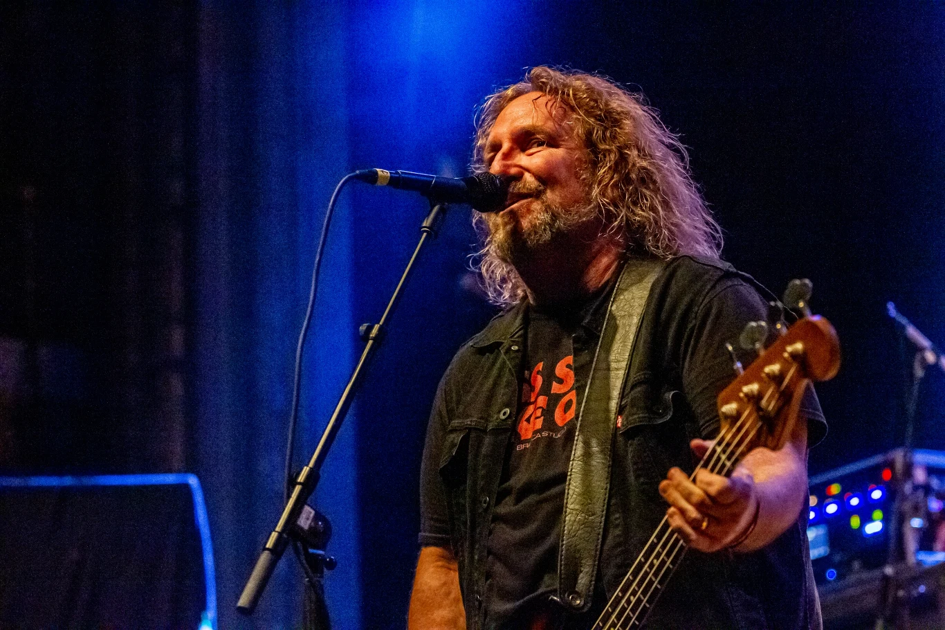 Mężczyzna z długimi, kręconymi włosami gra na gitarze basowej i śpiewa do mikrofonu na scenie koncertowej. Za nim widoczny niebieski reflektor sceniczny oraz sprzęt muzyczny.