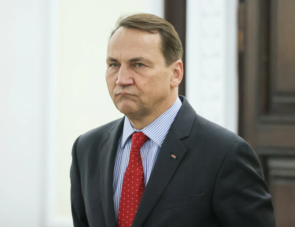 Radosław Sikorski zabrał głos w sprawie planu pokojowego dla Ukrainy Mężczyzna w średnim wieku w garniturze, niebieskiej koszuli w paski i czerwonym krawacie w białe kropki, stoi w formalnym otoczeniu, patrząc przed siebie z poważnym wyrazem twarzy.