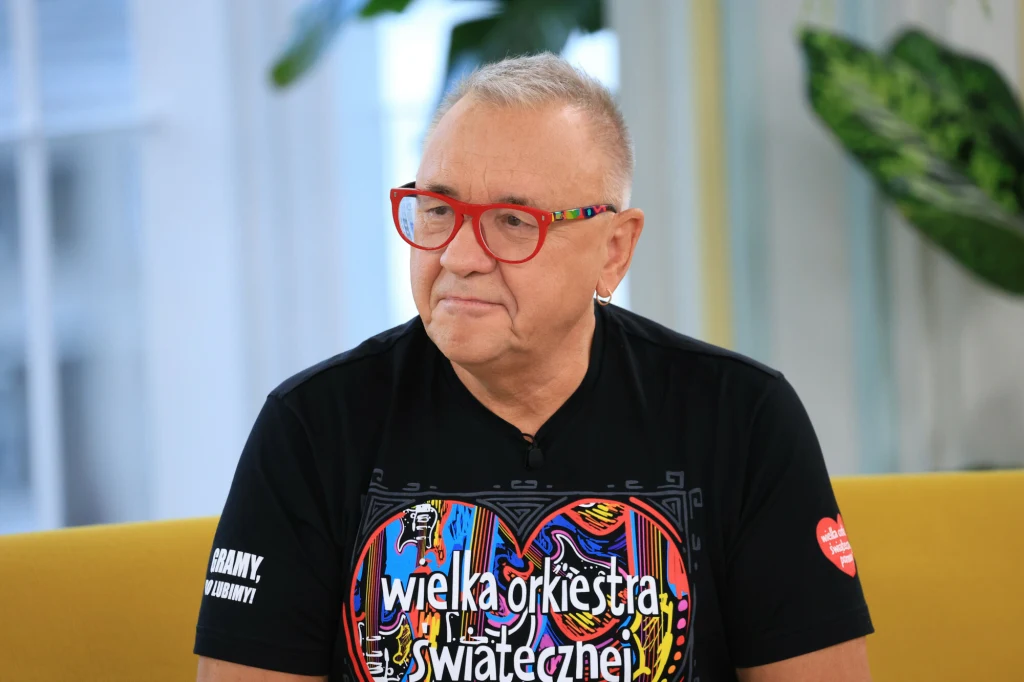 Mężczyzna w okularach z czerwonymi oprawkami siedzi na żółtej sofie, nosi czarną koszulkę z kolorowym wzorem oraz napisem 'wielka orkiestra świątecznej', w tle zielone rośliny doniczkowe.