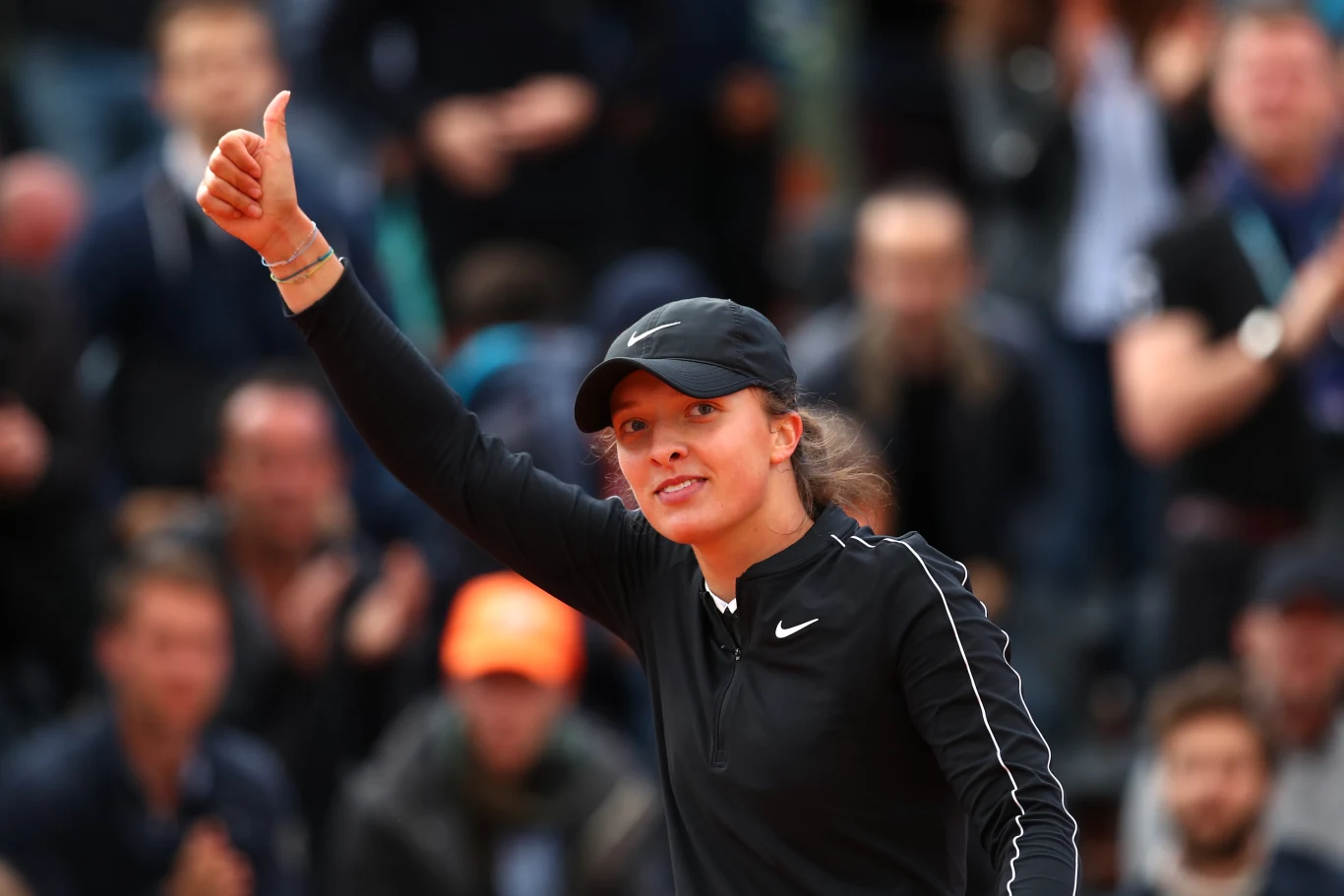 Iga Świątek Kobieta ubrana w czarny sportowy strój oraz czapkę z daszkiem unosi kciuk do góry, stojąc na tle zamazanej publiczności.