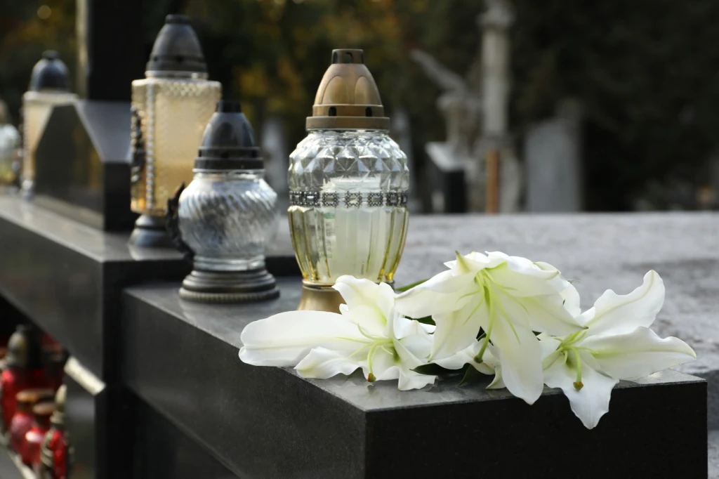 Białe lilie oraz kilka zniczy ustawionych na granitowym nagrobku, w tle rozmyte elementy cmentarza.