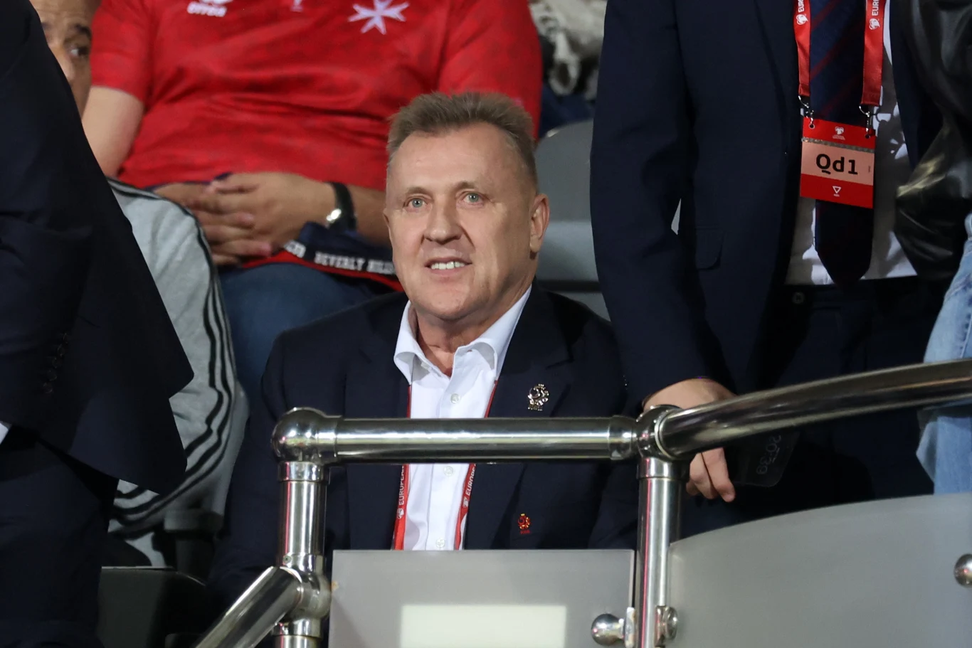 Cezary Kulesza Mężczyzna w garniturze siedzi na trybunach stadionu podczas wydarzenia sportowego, wokół znajdują się inne osoby, niektórzy ubrani w oficjalne identyfikatory i ubrania drużyny.