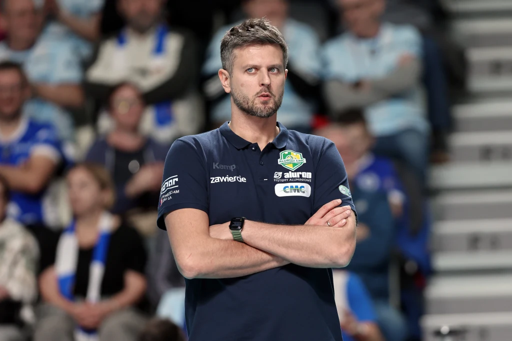 Michał Winiarski Mężczyzna w granatowej koszulce z logo sponsorów i skrzyżowanymi ramionami stoi na tle rozmytej publiczności, wyrażając skupienie i zaangażowanie.