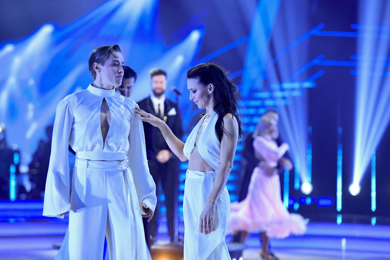 Katarzyna Zillmann, Janja Lesar Para tancerzy ubrana w eleganckie, białe stroje znajduje się na oświetlonej scenie podczas występu tanecznego, w tle widać innych uczestników oraz dynamiczne niebieskie światła.