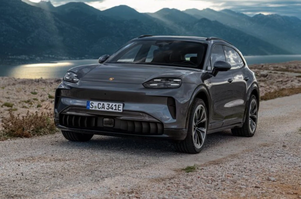 Szary nowoczesny samochód terenowy marki Porsche zaparkowany na żwirowej drodze, w tle widoczne są góry oraz jezioro oświetlone promieniami słońca, co tworzy malowniczy krajobraz.