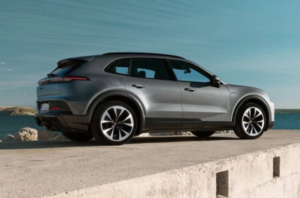 Srebrny SUV klasy premium stoi zaparkowany na betonowej powierzchni z widokiem na wodę i jasne niebo w tle. Auto wyróżnia się dużymi, sportowymi felgami, smukłą sylwetką oraz nowoczesnym designem świateł tylnych.