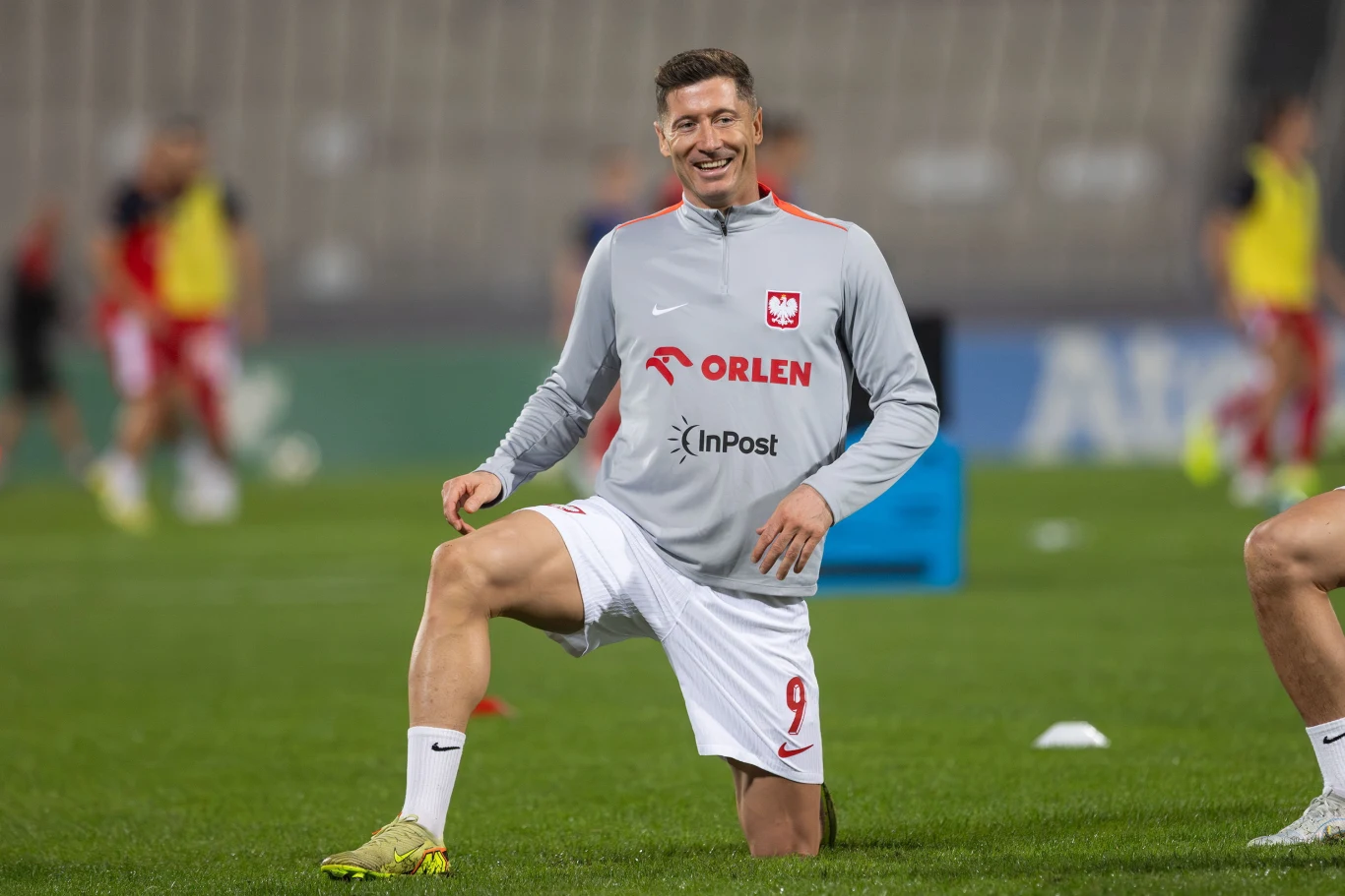 Robert Lewandowski Piłkarz w stroju reprezentacji Polski wykonuje rozgrzewkę na boisku, uśmiechając się, w tle widoczni inni zawodnicy trenujący.