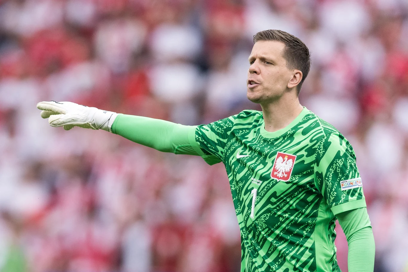 Wojciech Szczęsny Bramkarz ubrany w zielony strój z orłem na piersi stoi na boisku podczas meczu piłkarskiego, gestykulując w stronę kolegów z drużyny na tle rozmytej, dopingującej publiczności.