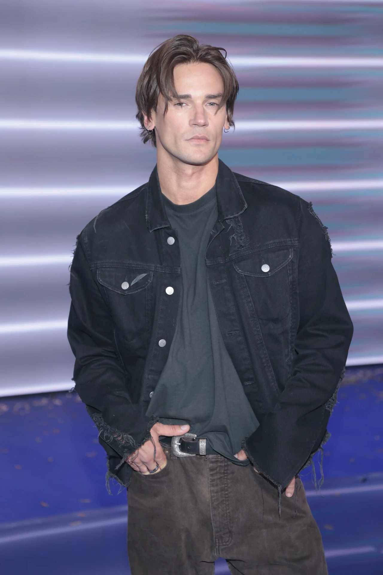 Mężczyzna z jasnymi włosami ubrany w czarną kurtkę jeansową oraz ciemny t-shirt i spodnie, stojący na tle z niebieskimi i fioletowymi pasami świetlnymi.