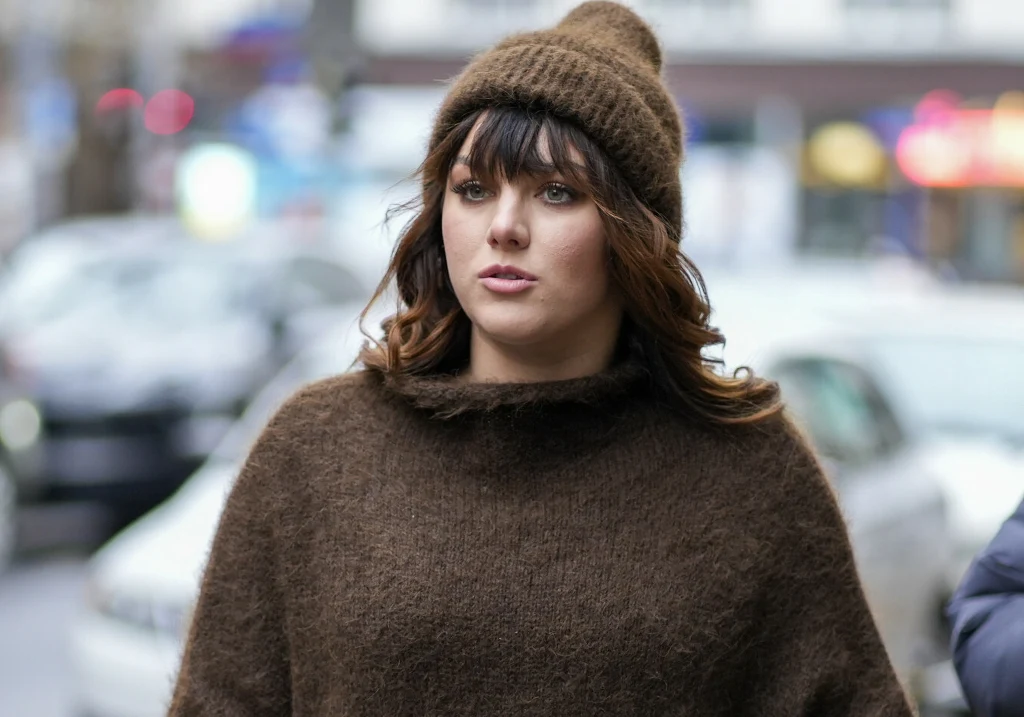 Ewa Farna Młoda kobieta ubrana w brązowy sweter i czapkę idąca po ulicy miasta, w tle rozmyte samochody oraz elementy miejskiego krajobrazu.