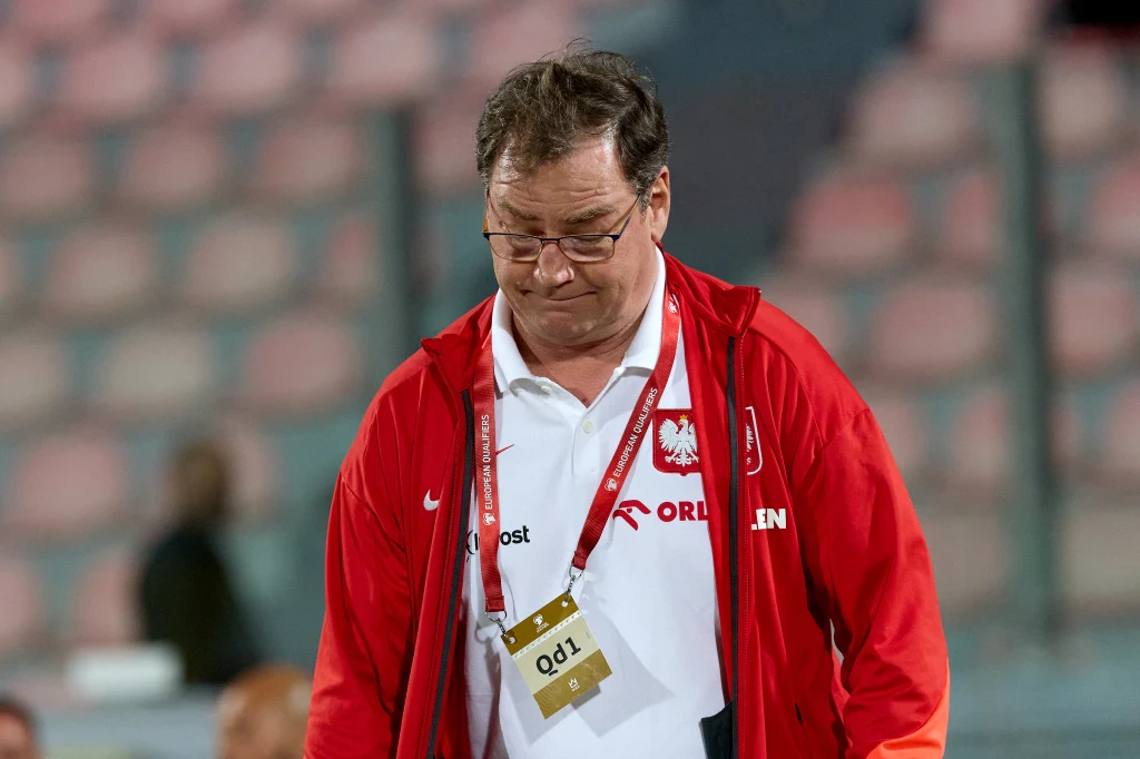 Jan Urban Mężczyzna w czerwonej kurtce z logiem Orlen oraz emblematem polskiej reprezentacji sportowej, pochylony, z identyfikatorem na szyi, w tle rozmyte trybuny stadionu.