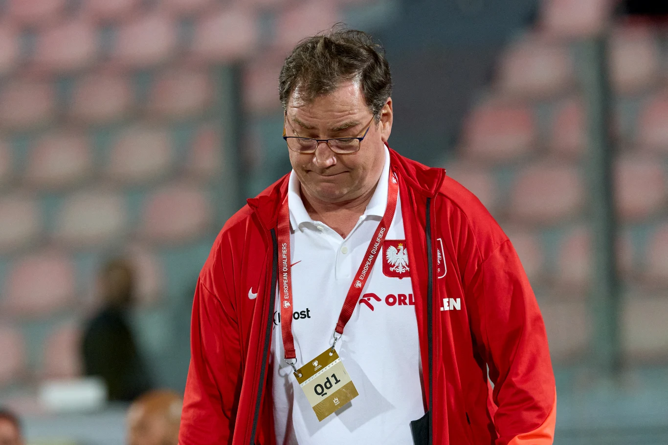 Mężczyzna w czerwonej kurtce z logiem Orlen oraz emblematem polskiej reprezentacji sportowej, pochylony, z identyfikatorem na szyi, w tle rozmyte trybuny stadionu.
