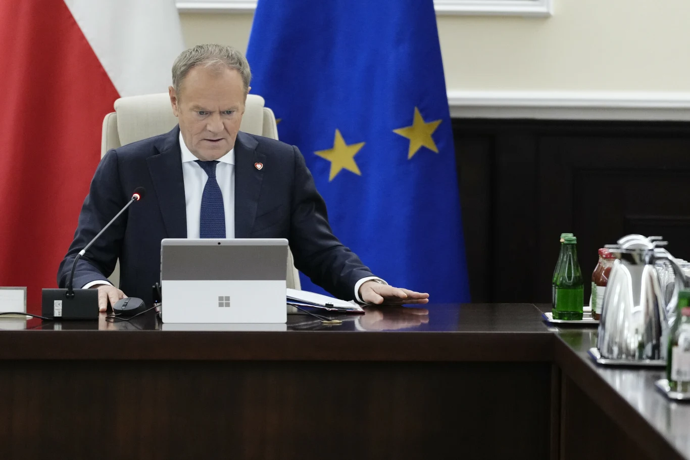 Donald Tusk pisze do prezydenta i opozycji Mężczyzna w garniturze siedzi przy stole konferencyjnym, patrząc w ekran laptopa. Na stole znajdują się karafka z wodą, szklanki oraz butelka z wodą mineralną. W tle widoczne są dwie flagi – Polski oraz Unii Europejskiej.